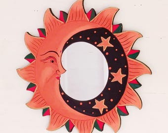 May include: A wooden sun and moon mirror with a crescent moon shape and a face on the sun. The moon is black with white stars and white dots. The sun is orange with green and red accents.