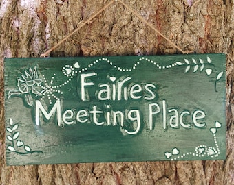 May include: A rectangular, dark green wooden sign with the words "Fairies Meeting Place" in white, whimsical lettering. The sign is decorated with white floral and dot accents and is suspended by a natural twine string.