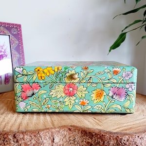 May include: A rectangular, hand-painted wooden box with a light teal background and floral design. The flowers are in shades of pink, yellow, and orange, with green leaves and vines. The box has a small, round, gold-colored knob.