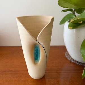 May include: A beige ceramic vase with a distinctive folded design. It has a teal accent and a black line detail. The vase is on a wooden surface, with a plant in a white pot in the background.