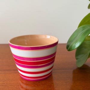 May include: A striped ceramic plant pot with bands of white, pink, and red. The interior of the pot is a light peach colour. The pot is displayed on a wooden surface.
