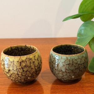 May include: Two small, round ceramic bowls with a textured, crackled glaze. One bowl is yellow-brown, the other is green-brown. The bowls are on a wooden surface, with green plant leaves in the background.