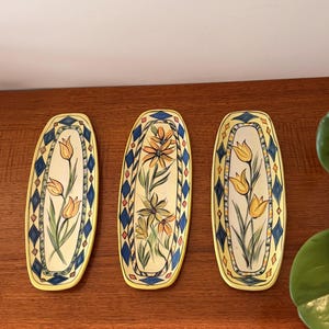May include: Three oblong ceramic serving dishes with floral designs. Each dish features a yellow background with a painted flower and green stems. The edges are decorated with blue diamond patterns and yellow borders.