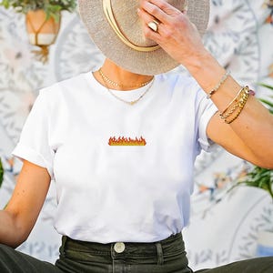 Peut inclure: T-shirt blanc avec un petit motif de flamme brod&eacute;e en nuances d'orange et de jaune. Le t-shirt a des manches courtes et un col rond. Le motif est centr&eacute; sur la poitrine. Le t-shirt est port&eacute; avec un pantalon vert olive.