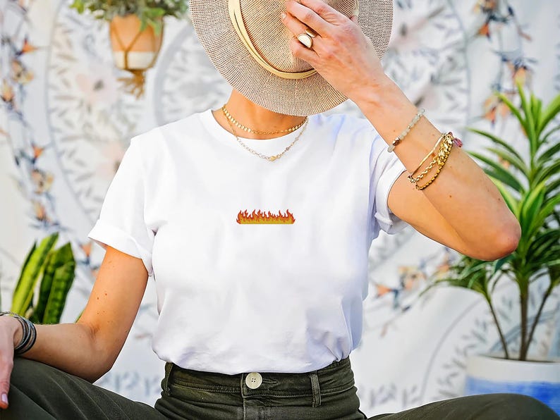 Peut inclure: T-shirt blanc avec un petit motif de flamme brod&eacute;e en nuances d'orange et de jaune. Le t-shirt a des manches courtes et un col rond. Le motif est centr&eacute; sur la poitrine. Le t-shirt est port&eacute; avec un pantalon vert olive.