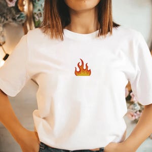 Peut inclure: T-shirt blanc avec un petit motif de flamme brod&eacute;e en rouge, orange et jaune. Le haut a un col rond et des manches courtes. Le motif de la flamme est centr&eacute; sur la poitrine.