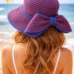 Può includere: Un cappello da sole all'uncinetto viola con un grande dettaglio a fiocco. Il cappello presenta un'ampia tesa e un bordo blu a contrasto. La persona indossa il cappello con lunghi capelli rossi ondulati, con una spiaggia e un oceano sullo sfondo.