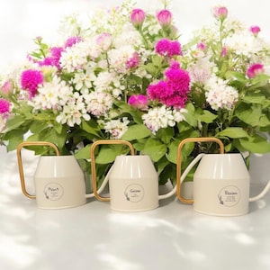 May include: Three white watering cans with gold handles and spouts. Each can has a different word on it: "Plant", "Grow", and "Bloom". The cans are sitting on a white surface in front of a bouquet of white and pink flowers.