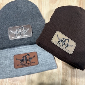 May include: Three knitted beanies in grey, brown, and light grey. Each beanie features a rectangular leather patch with the text "CTT Oil Exploration and Cattle Midland, Texas" and an oil derrick graphic.