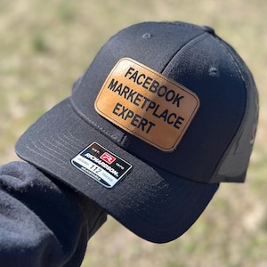 Pode incluir: Boné de caminhoneiro preto com um remendo de couro marrom que diz "Facebook Marketplace Expert". O boné é um Richardson 112 Trucker.