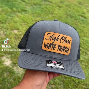 May include: Gray and black trucker hat with a leather patch that reads "High Class WHITE TRASH" in a cursive font. The hat has a Richardson 112 tag.