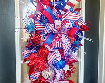May include: A patriotic teardrop-shaped door swag featuring red, white, and blue ribbons, metallic ornaments, and glittery star accents. The arrangement includes striped and star-patterned fabric ribbons, shiny red and blue spheres, and thin wire sprays.