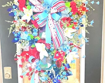 May include: A patriotic door wreath with red, white, and blue ribbons, bows, and floral accents. The wreath features a mix of artificial flowers and greenery, creating a festive and decorative display for the front door.