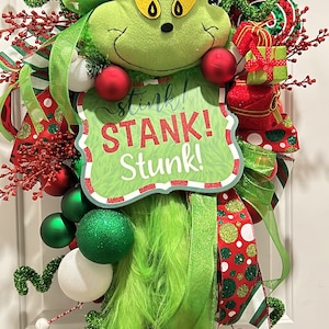 May include: A whimsical Christmas wreath featuring a plush Grinch head wearing a red Santa hat. The wreath includes green and red ornaments, ribbons, and a sign that reads "Stink! Stank! Stunk!"