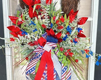 May include: A patriotic wreath with a grapevine base, decorated with red, white, and blue flowers and ribbons. The wreath includes a bow made from ribbon with an American flag design, suitable for door decoration.