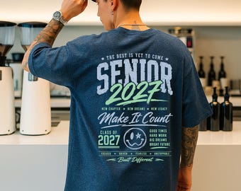 May include: A navy blue t-shirt with the text "SENIOR 2027" in white and green. The shirt also features the phrases "Make It Count", "New Chapter, New Dreams, New Legacy", and "Built Different". The person in the image is wearing a navy blue baseball cap.