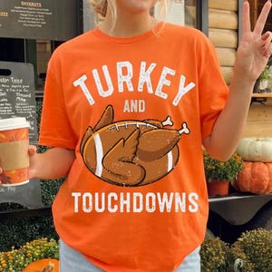 May include: An orange short-sleeve t-shirt featuring a graphic of a brown roasted turkey shaped like an American football with white laces. The text above the graphic reads TURKEY AND and the text below reads TOUCHDOWNS in white letters.