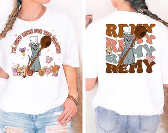 Può includere: T-shirt bianca con un topo chef dei cartoni animati che tiene un cucchiaio di legno, circondato da snack colorati. La parte anteriore dice "I'M JUST HERE FOR THE SNACKS". La parte posteriore della maglietta dice "Remy" in un font retrò.