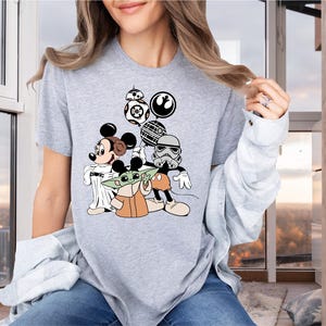 May include: A gray t-shirt with a graphic print of Mickey Mouse, Yoda, and a Stormtrooper from Star Wars. The characters are holding a Death Star and a lightsaber.