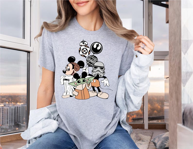 May include: A gray t-shirt with a graphic print of Mickey Mouse, Yoda, and a Stormtrooper from Star Wars. The characters are holding a Death Star and a lightsaber.