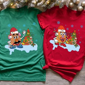 Könnte beinhalten: Zwei T-Shirts, eines grün und eines rot, jeweils mit einer Cartoon-Kartoffel-Figur mit Weihnachtsmütze, Lichterketten, Schneeflocken und Geschenken. Die Kartoffel hat einen schwarzen Schnurrbart und steht auf einer blauen Wolke.