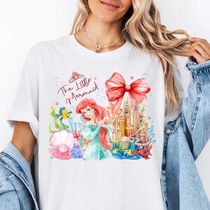 May include: White t-shirt featuring a colorful illustration of Ariel from The Little Mermaid. The design includes a red bow, a castle, and the text "The Little Mermaid" in red script. The artwork is a watercolor style.