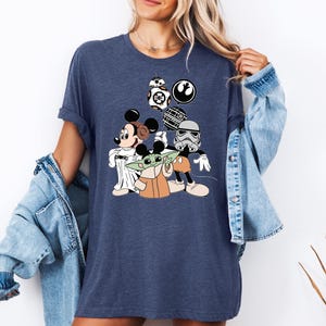 May include: A navy blue t-shirt with a graphic of Mickey Mouse, Yoda, and Stormtrooper characters from Star Wars. The characters are wearing Mickey Mouse ears and holding Star Wars themed items.