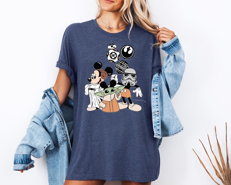 May include: A navy blue t-shirt with a graphic of Mickey Mouse, Yoda, and Stormtrooper characters from Star Wars. The characters are wearing Mickey Mouse ears and holding Star Wars themed items.