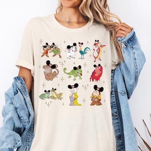 Op de afbeelding: Crèmekleurig T-shirt met cartoonfiguren met Mickey Mouse oren. Het shirt toont verschillende personages zoals een aap, een kameleon en een haan, allemaal versierd met de iconische oren. Kleine sterrenafbeeldingen zijn verspreid rond de personages.