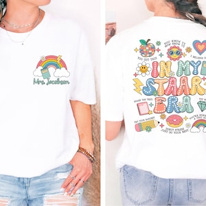 Op de afbeelding: Wit T-shirt met een regenboog-, boek- en wolkenontwerp aan de voorkant en een kleurrijke afbeelding met de tekst "In My Staar Era" aan de achterkant. De afbeelding bevat een donut, potlood, bliksem en andere schoolthema-pictogrammen.