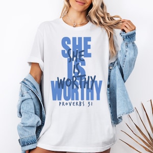 Può includere: T-shirt bianca con la scritta blu "SHE IS WORTHY". Sotto è stampato "PROVERBI 31". La maglietta fa parte di una collezione di abbigliamento. Il testo è in grassetto.