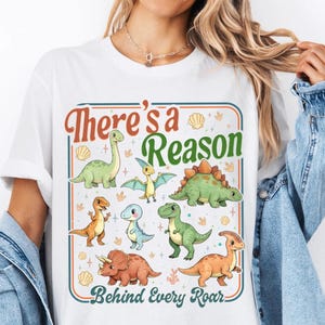 Könnte beinhalten: Weißes T-Shirt mit einem bunten Dinosaurier-Motiv und dem Text "There's a Reason Behind Every Roar". Das Design zeigt verschiedene Cartoon-Dinosaurier in Grün, Orange und Blau, eingebettet in einen dekorativen Rahmen.