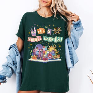 May include: Dark green t-shirt featuring the text "it's a small world!" above a colorful illustration of a storybook scene with buildings, characters, and a sun. The shirt has short sleeves and a relaxed fit.