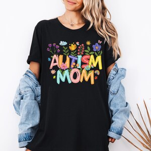 Op de afbeelding: Zwart t-shirt met de tekst "Autism Mom" in kleurrijke, speelse letters, omringd door bloemen- en regenboogafbeeldingen. Een casual kledingstuk voor dagelijks gebruik.