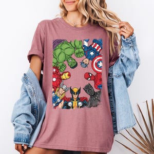 A woman wearing a pink t-shirt with a graphic of various Marvel superheroes, including Hulk, Captain America, Spider-Man, and others, standing together.