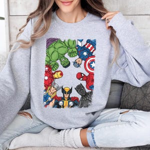 A woman wearing a grey sweatshirt with a graphic of various Marvel superheroes, including Hulk, Captain America, Spider-Man, and others, sitting on a couch.