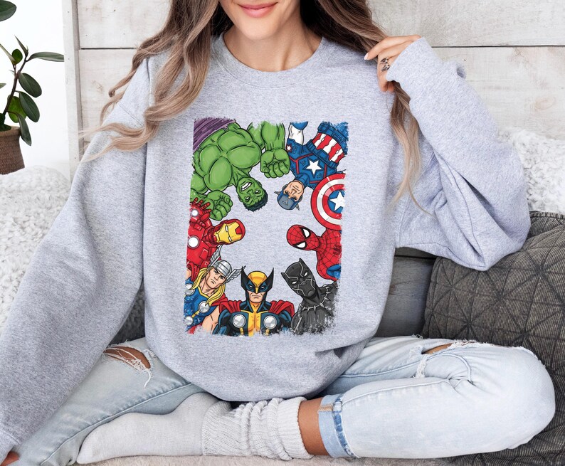 A woman wearing a grey sweatshirt with a graphic of various Marvel superheroes, including Hulk, Captain America, Spider-Man, and others, sitting on a couch.