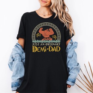 May include: Black t-shirt with a vintage-style graphic of a cartoon character with the text "Just an Ordinary Demi-Dad".