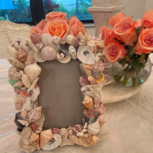 May include: A picture frame made entirely of seashells. The frame is a light brown color and has a variety of different seashells, including clams, oysters, and snails. The frame is sitting on a white table.