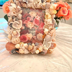 May include: A picture frame made entirely of seashells. The frame is decorated with a variety of seashells in different shapes, sizes, and colors. The frame is holding a picture of a floral arrangement.