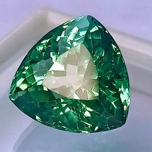 May include: A faceted, triangular gemstone with a vibrant green hue. The gem's surface reflects light, showcasing a complex internal structure. The shape is a perfect equilateral triangle, ideal for jewelry or collection.