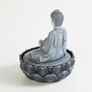 May include: A gray Buddha statue seated in a lotus flower base. The statue is in a meditative pose, holding a lotus flower. The base is intricately detailed with a floral pattern. The statue is made of a stone-like material.