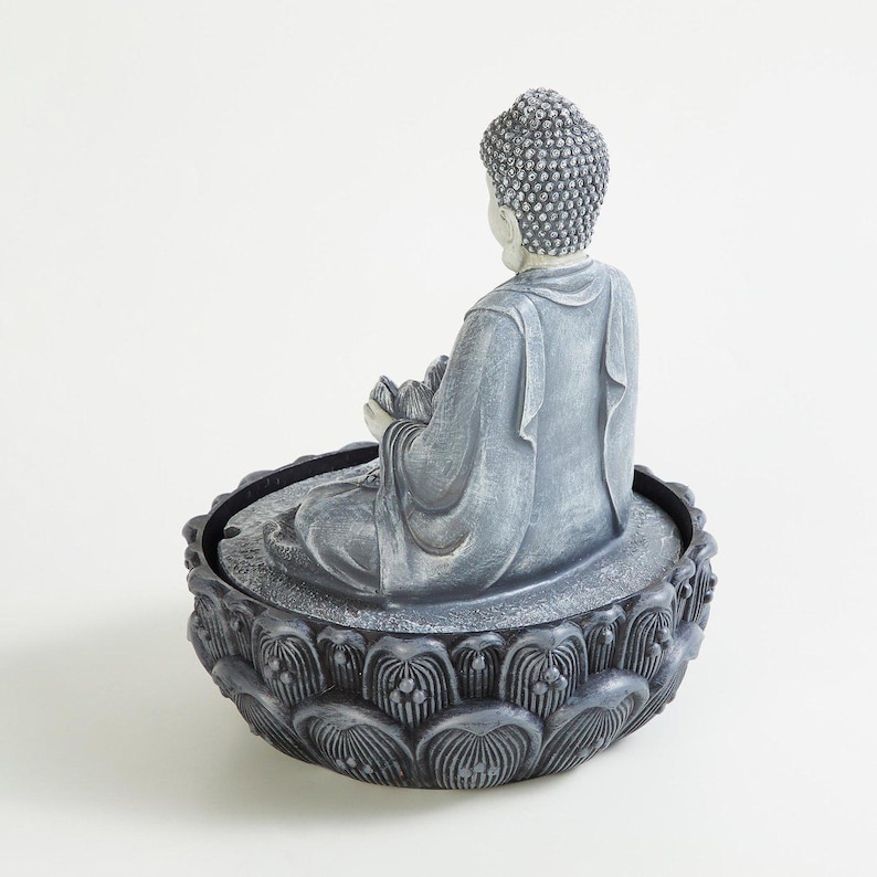 May include: A gray Buddha statue seated in a lotus flower base. The statue is in a meditative pose, holding a lotus flower. The base is intricately detailed with a floral pattern. The statue is made of a stone-like material.