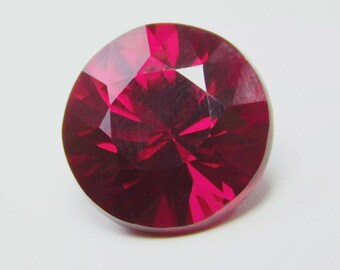 May include: A faceted, round-cut ruby gemstone. The gem is a deep, saturated red color with multiple facets that reflect light. The gemstone is set against a white background.
