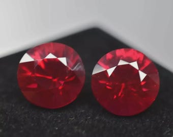 May include: Two round, faceted, deep red gemstones. Each gem has a series of triangular facets at the top, creating a starburst effect. The gems are set against a dark background, highlighting their vibrant color and brilliance.