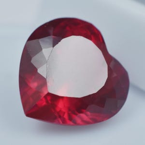 May include: A heart-shaped gemstone with a deep red colour and multiple facets. The gem features a large, unpolished, white area in the centre, surrounded by the polished, reflective facets. This gemstone is likely used in jewellery.