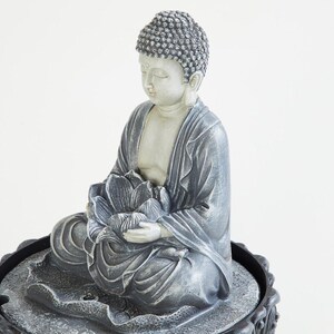 May include: A seated Buddha statue in a meditative pose, holding a lotus flower. The statue is primarily gray with a light beige face and hands. The Buddha is seated on a decorative base, creating a serene and spiritual ambiance.