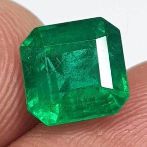 Puede incluir: Una vibrante piedra preciosa verde con talla esmeralda. La gema tiene forma rectangular con bordes biselados, mostrando un color verde intenso y profundo. Las facetas reflejan la luz, resaltando la claridad y el brillo de la piedra. La gema es sostenida por los dedos de una persona.