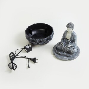May include: A gray resin Buddha statue seated on a lotus flower base, holding a lotus flower. Includes a black basin with decorative edges, a small pump, and a power cord. The fountain is designed for indoor use.