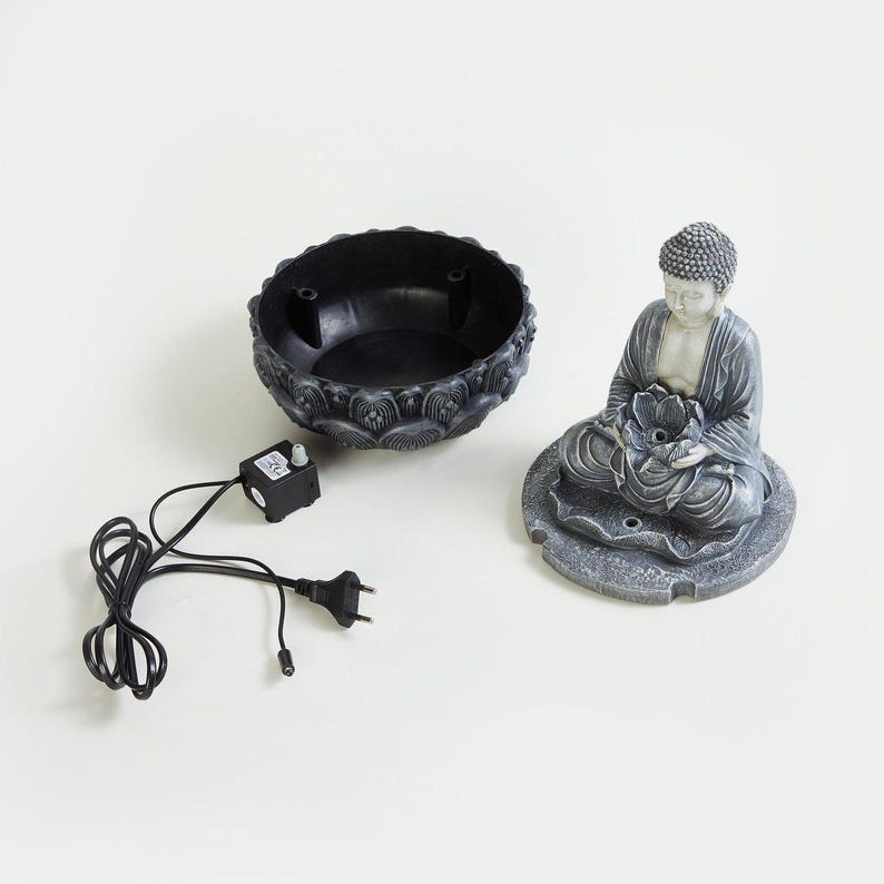 May include: A gray resin Buddha statue seated on a lotus flower base, holding a lotus flower. Includes a black basin with decorative edges, a small pump, and a power cord. The fountain is designed for indoor use.